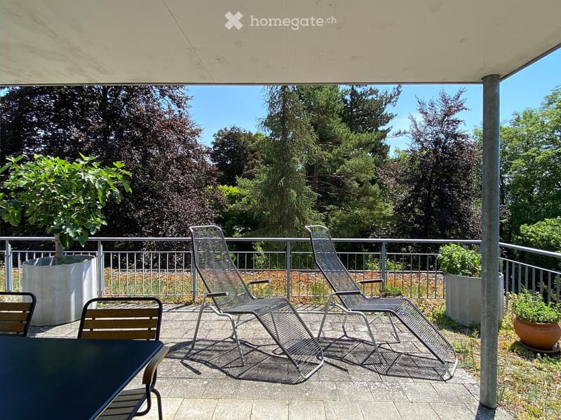 Balcony with a table, chairs, and lounge chairs, surrounded by greenery and a metal railing.