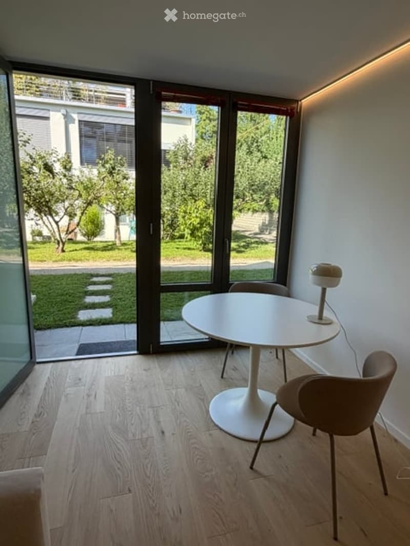 Modern, minimalist dining area with a white round table, two chairs, and a small lamp on the table. Large glass doors open to a green outdoor space.