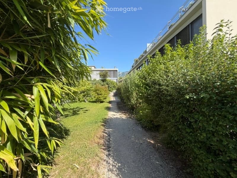 A well-maintained garden path lined with lush greenery leads to a modern building with a balcony and large windows.