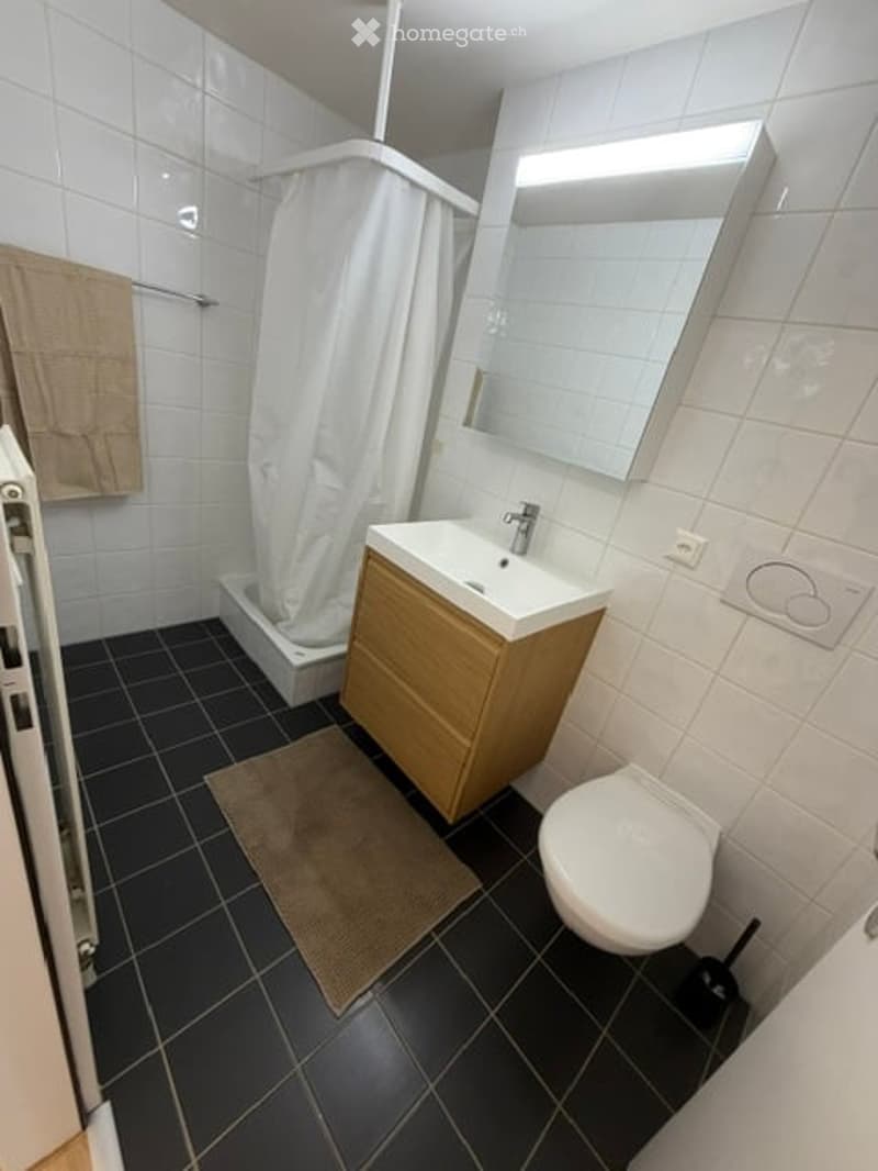 Modern bathroom with white tiles, black floor tiles, a wooden vanity cabinet, a sink, a mirror, a toilet, and a shower area with a white curtain.