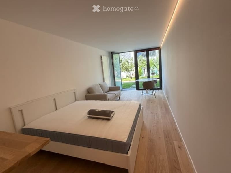 Modern, minimalist bedroom with a large bed, a sofa, a chair, and a small table near the balcony. The room features light wooden flooring and white walls.