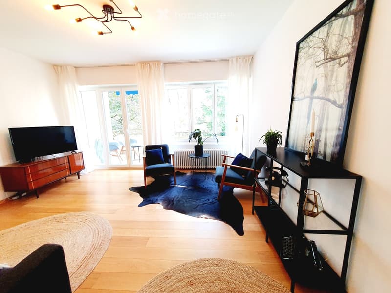 Bright living room with wooden flooring, large windows, and modern decor including a sofa, chairs, a coffee table, a television, and a large mirror.