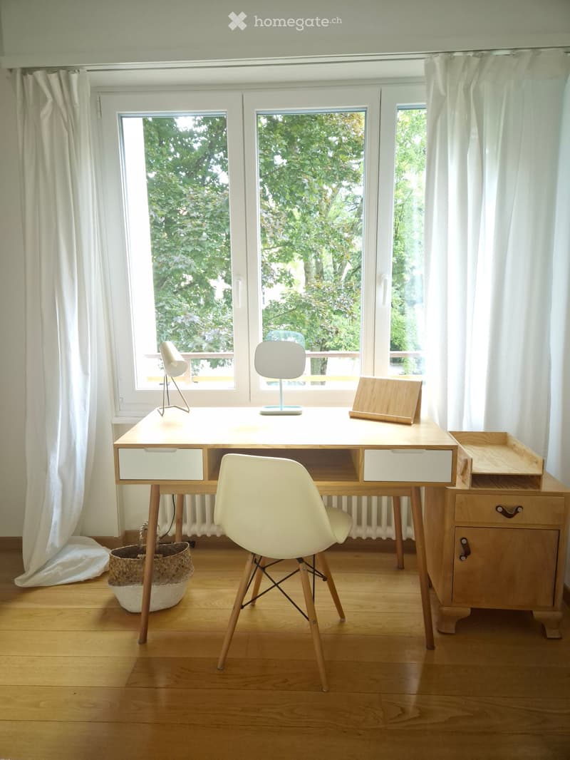Bright room with wooden floor, large windows, white desk, chair, lamp, and small basket.