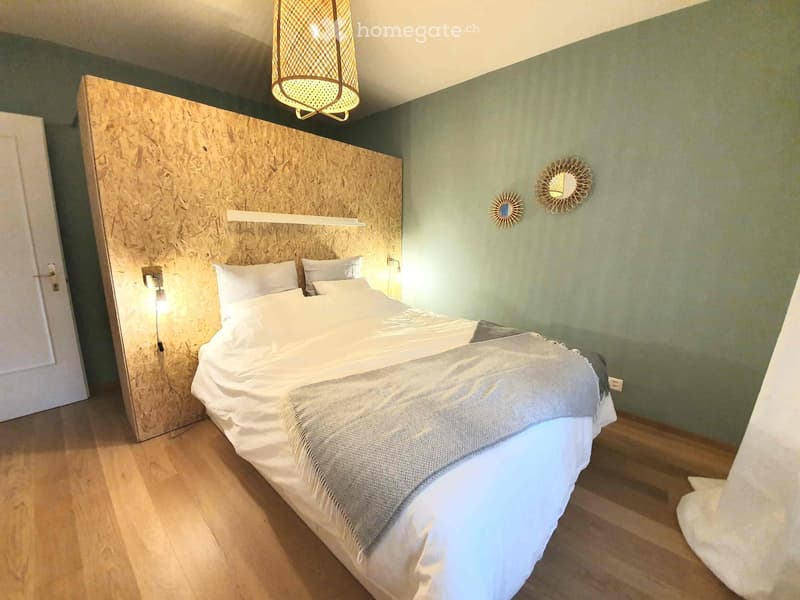 Modern bedroom with a large wooden headboard, a neatly made bed with white linens, a wall-mounted mirror, and a decorative lamp above the bed.