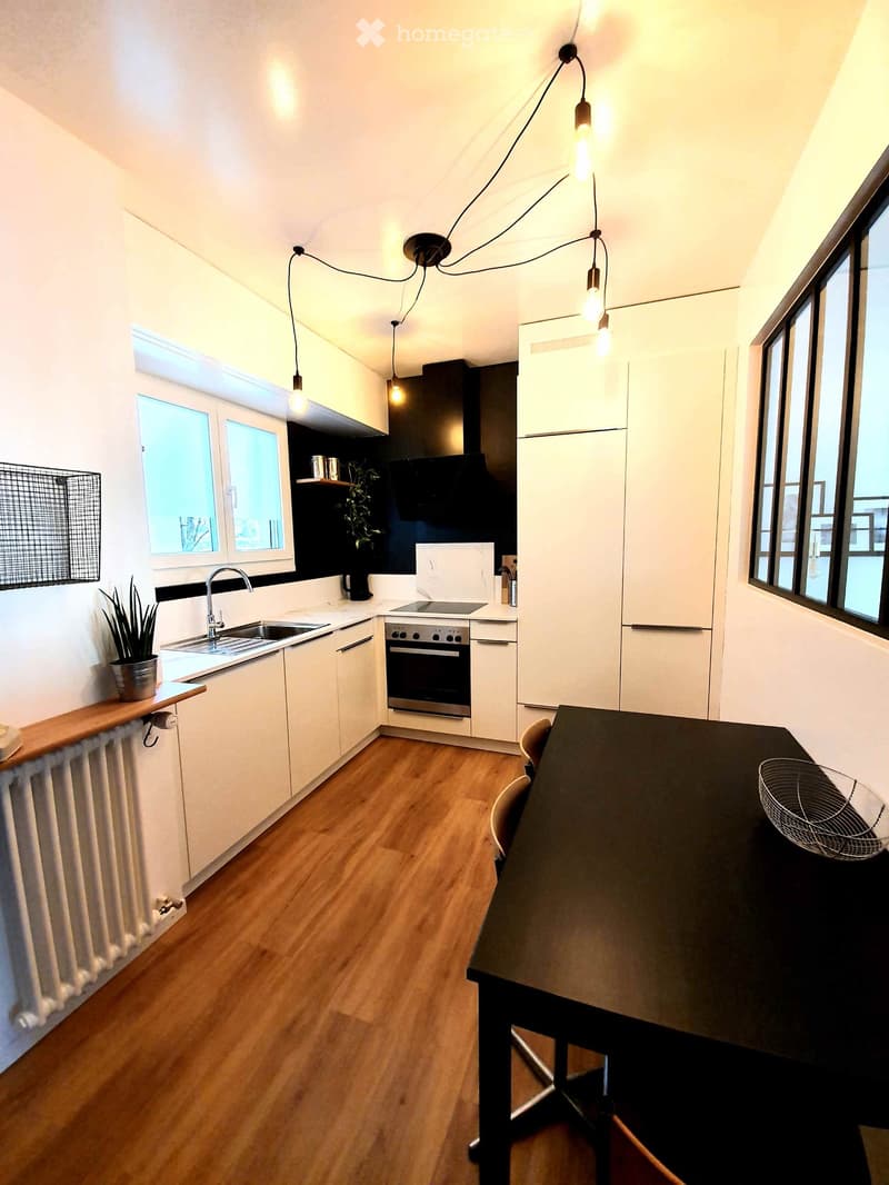 Modern kitchen with white cabinets, wooden flooring, stainless steel appliances, and pendant lighting. Features a sink, oven, refrigerator, and a small dining area with a table and chairs.