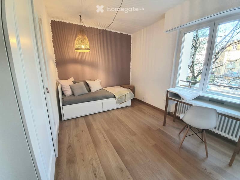 Cozy studio with a bed, desk, chair, and a hanging lamp. Large windows provide natural light.