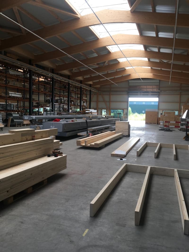 Large, empty warehouse space with exposed wooden beams and concrete floor, filled with stacks of wooden planks and construction materials.
