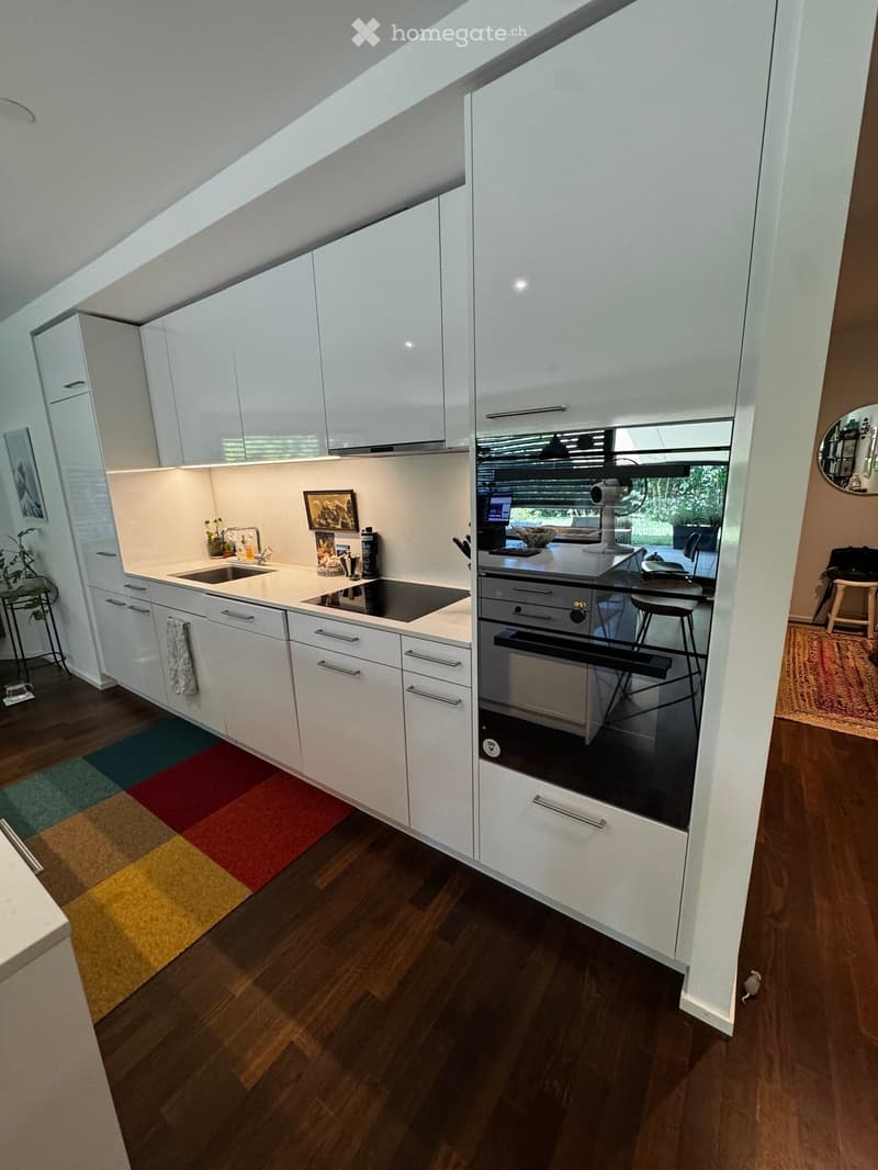 Modern kitchen with white cabinets, stainless steel appliances, and a wooden floor. Features a sink, stove, oven, and fridge, with a small plant and picture frame on the counter.