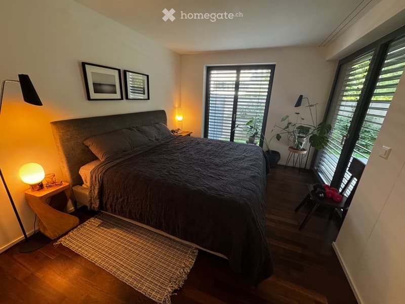 Modern bedroom with a large bed, nightstand, floor lamp, and large windows with blinds. Includes a small rug, a stool, and a plant near the window.