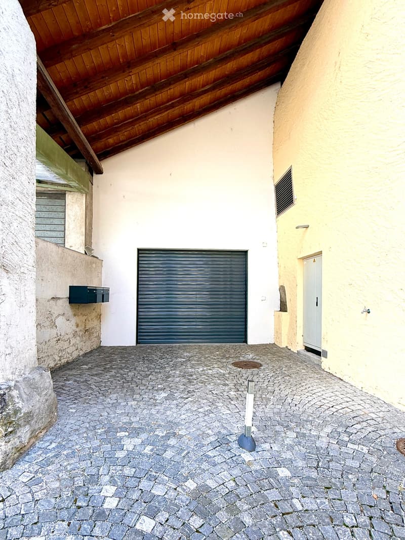 Narrow cobblestone pathway between two buildings, with a garage door, mailbox, and a small white post.