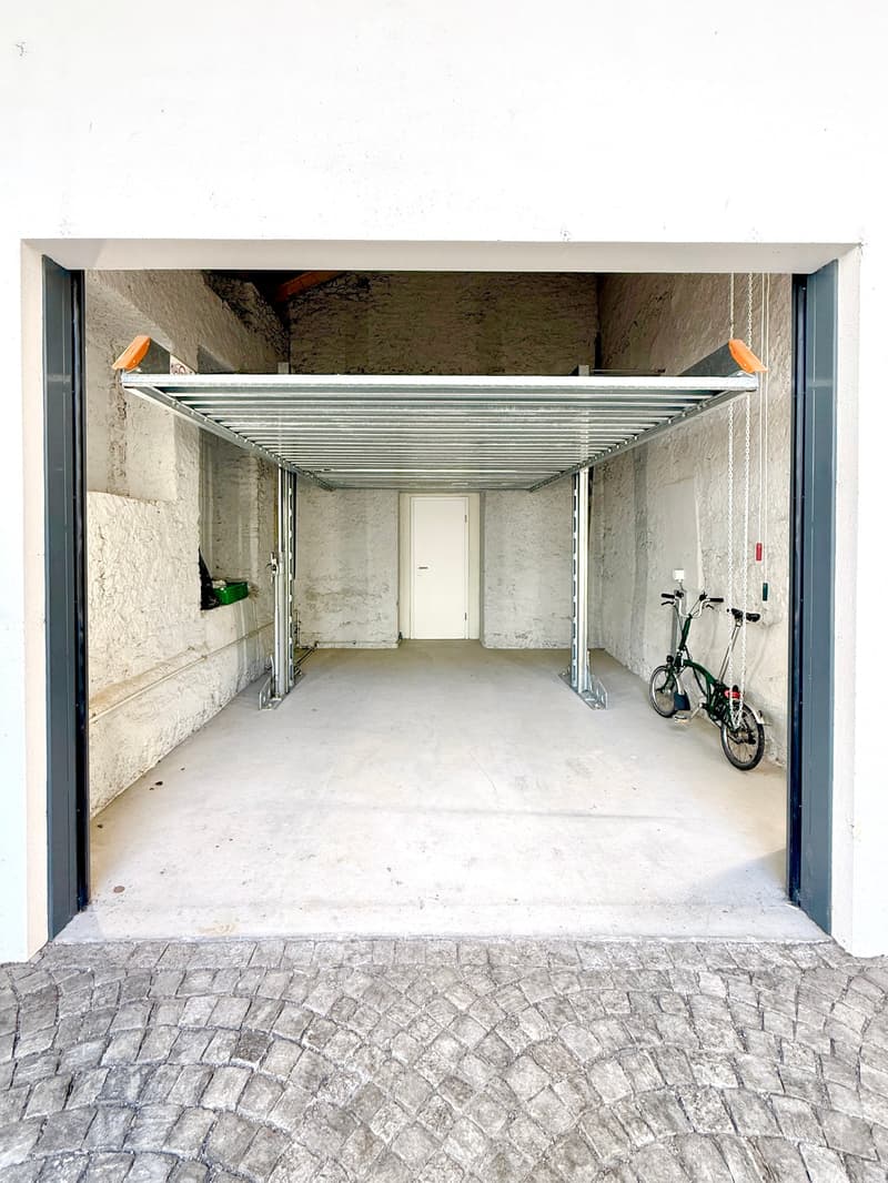 A covered, open-air corridor with concrete walls and a metal roof structure, featuring a bicycle parked on the right side and a white door at the far end.