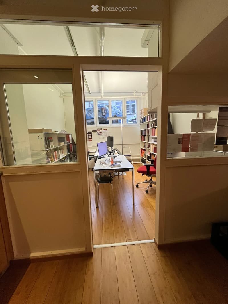 Open door with wooden floor, desk with monitor, red chair, book shelves, office supplies