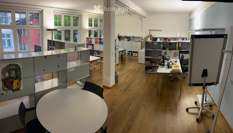 An empty office space with round table, several black chairs, cabinets, bookcases, and multiple computer equipment. There are windows with trees outside, and the room has a wooden floor.