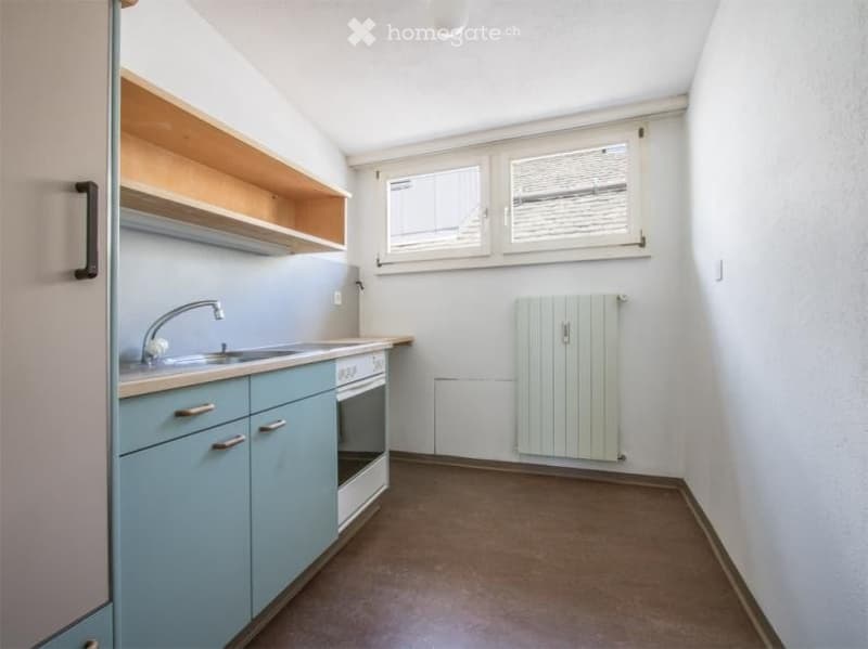 A small, modern kitchen with light blue cabinets, a sink, an oven, and built-in wooden shelves above the counter. The space features a radiator and two windows, with a neutral color palette and clean lines.