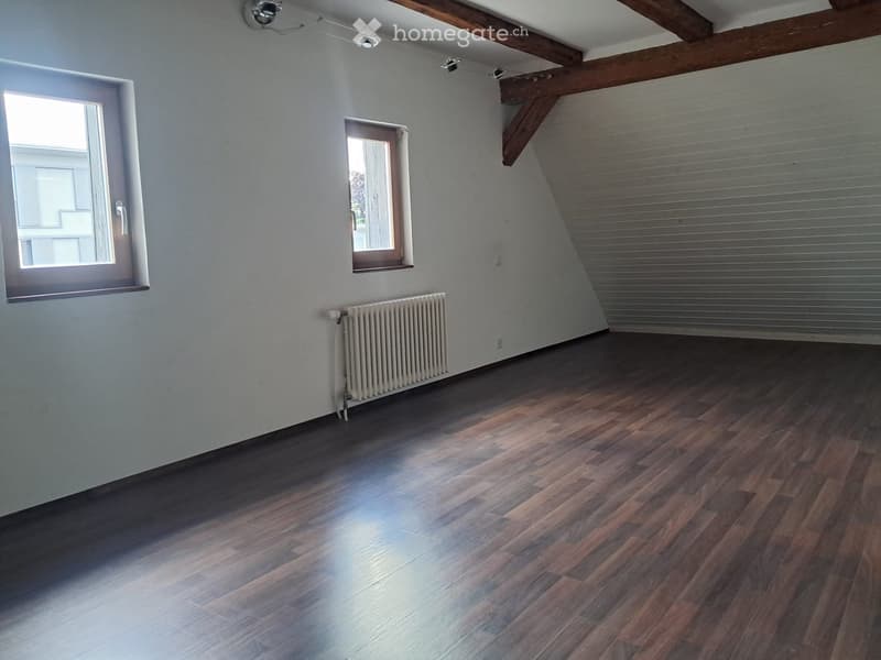 Empty room with wooden flooring, white walls, and exposed wooden beams on the ceiling. Features include a radiator, two windows, and a metal shutter on one wall.