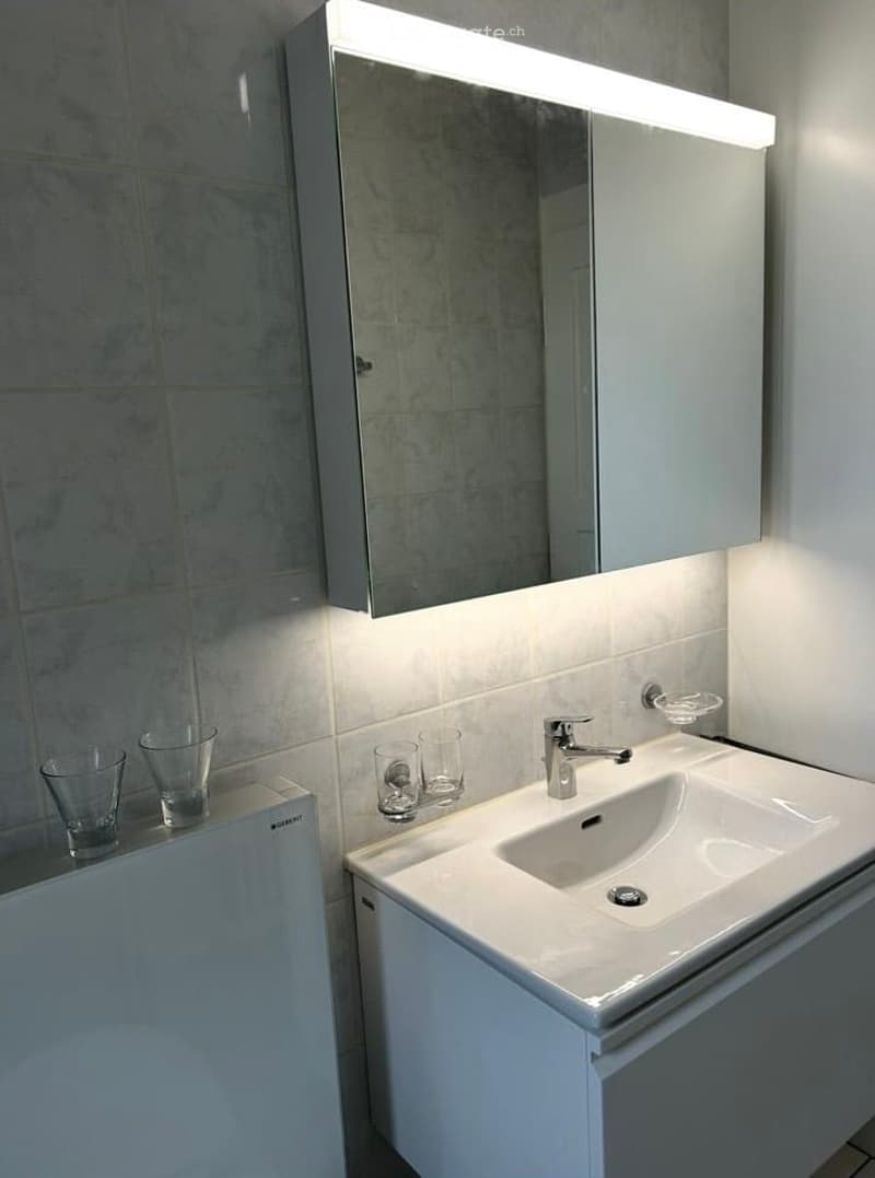 Modern bathroom with a white sink, chrome faucet, mirrored cabinet with built-in lighting, and several glass cups on the counter.