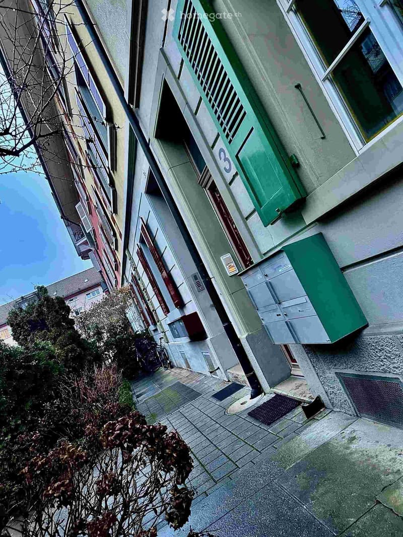 Multi-story residential building with green shutters, numbered entrance, and adjacent greenery.