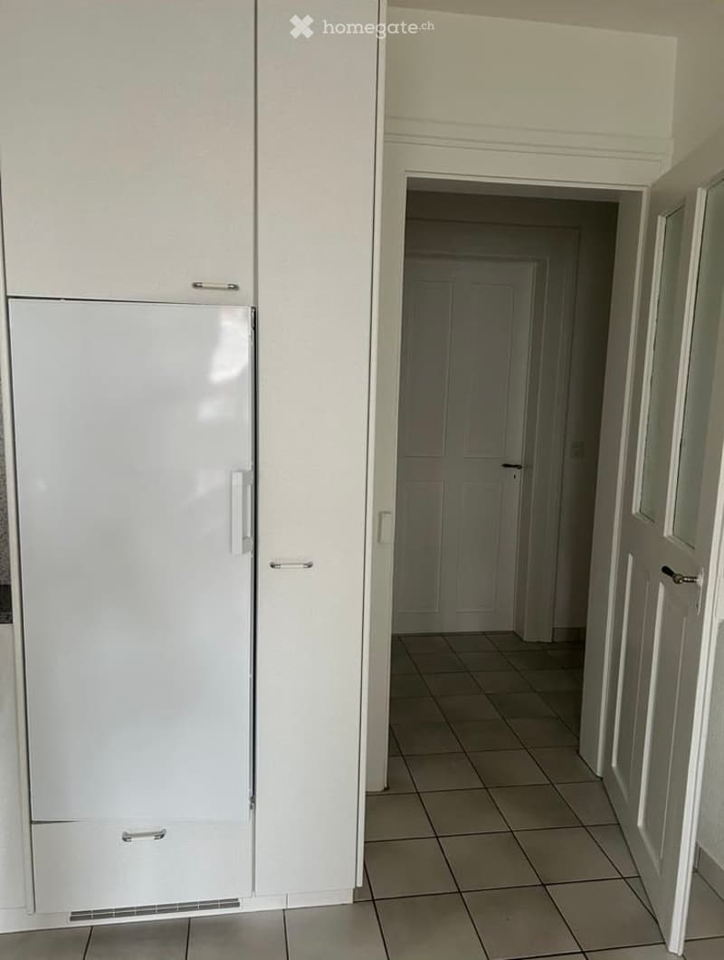 A modern kitchen with white cabinets and a large refrigerator, leading to a tiled hallway with doors.