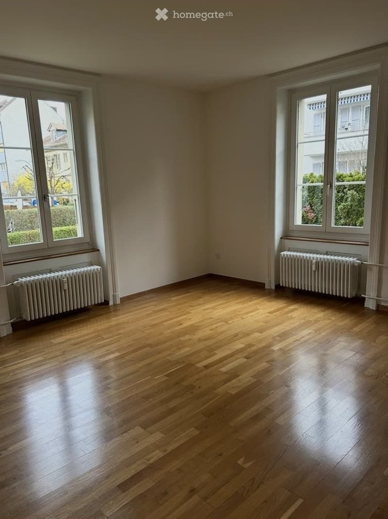 Empty room with wooden flooring, large windows, and radiators on both sides.