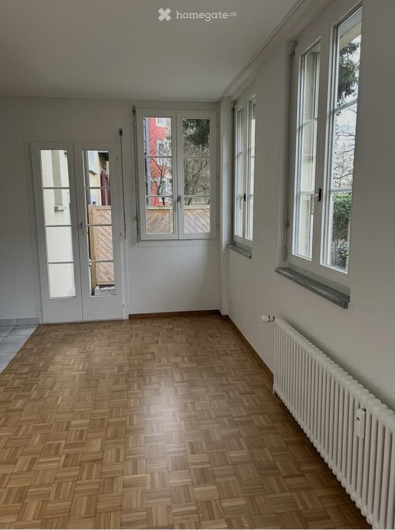 Empty room with wooden herringbone flooring, white walls, large windows, and a radiator. No furniture or equipment present.
