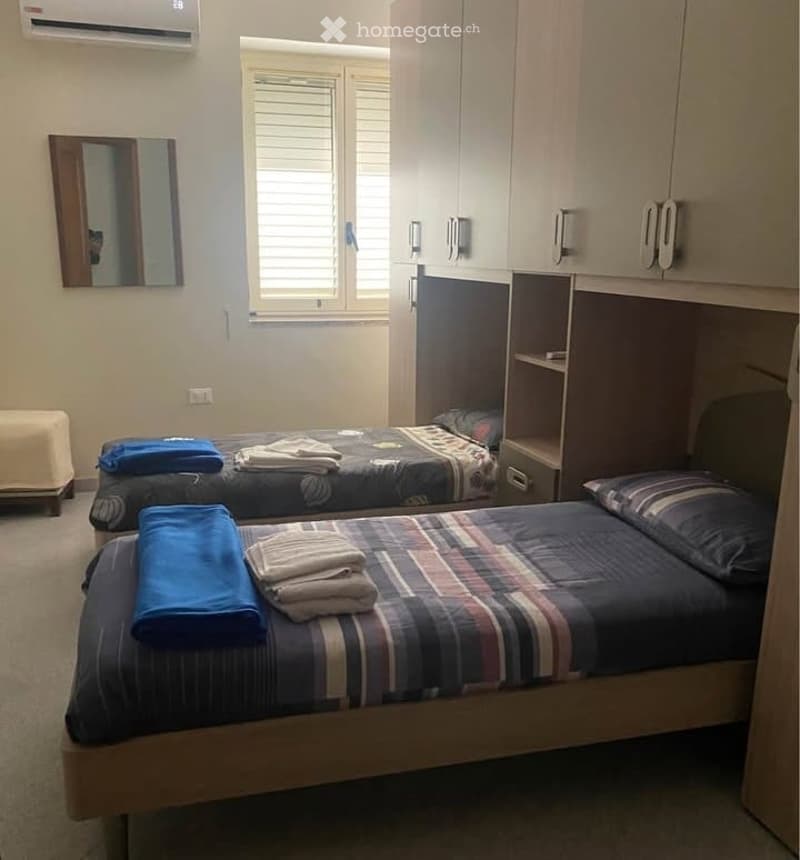Cozy bedroom with two single beds, neatly made with patterned bedspreads and folded towels, a mirror on the wall, built-in cabinets, and a small stool near the window.