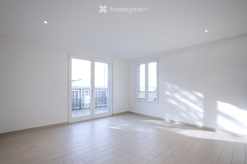 Empty room with white walls, wooden floor, large sliding glass door leading to a balcony, and a window with white frame.