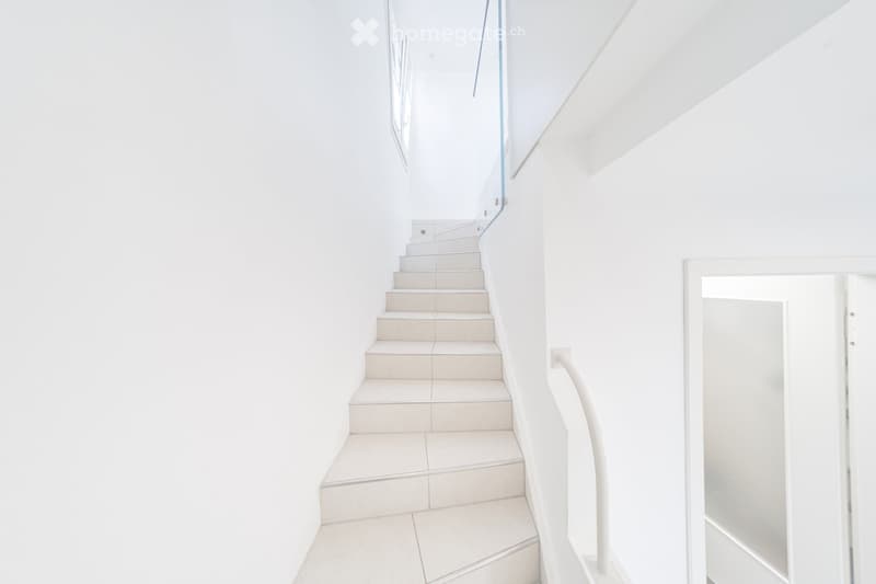 Modern, minimalist staircase with white tiled steps and a glass railing, leading upwards in a bright, open space.