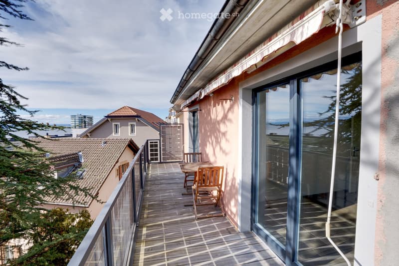 Balcony with wooden decking, glass railing, table and chairs, overlooking neighboring houses and a cityscape.