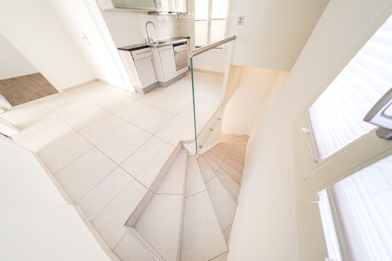 Modern hallway with tiled floor, white walls, staircase leading to an upper level, and a small kitchen area visible at the end.