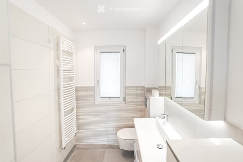 Modern bathroom with white tiles, a large mirror, a sink, a toilet, and a radiator. The space is bright with natural light from a window.