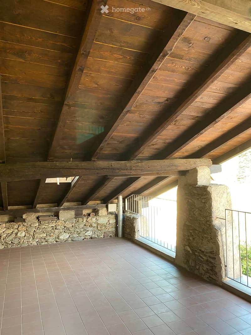 An unfurnished room with exposed wooden beams on the ceiling, stone walls, tiled floor, and large windows allowing natural light to enter.