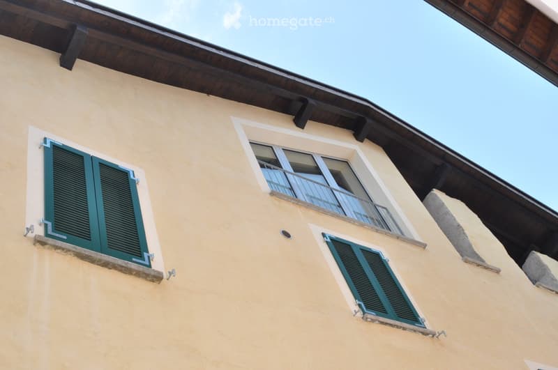 A section of a building with light yellow walls, green shutters, and a small balcony with a metal railing.