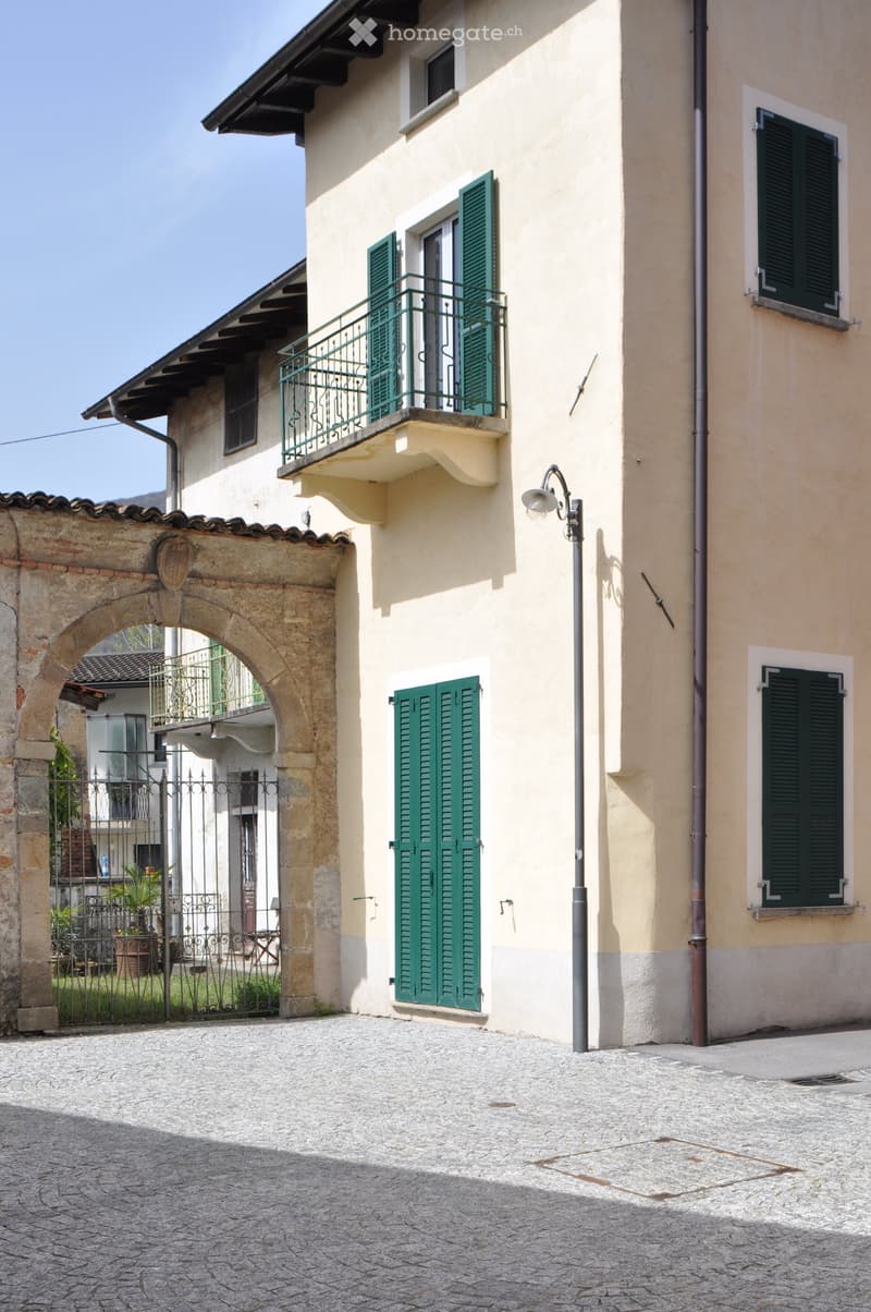 Two-story building with light yellow walls, green shutters, and a balcony with metal railing. The entrance features an arched stone doorway leading to a gated courtyard with greenery.