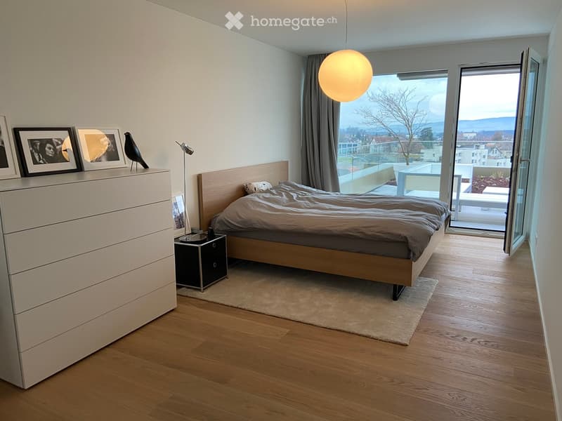 modern bedroom with wooden floor, large bed, white dresser, nightstand, lamp, picture frames, open balcony with view