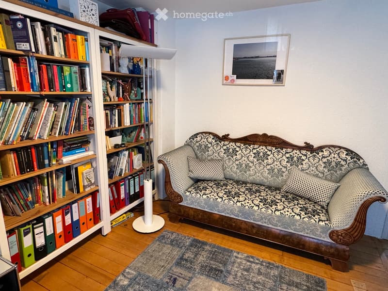 A cozy living room with a patterned sofa, wooden bookshelf filled with books and binders, a floor lamp, and a framed picture on the wall.