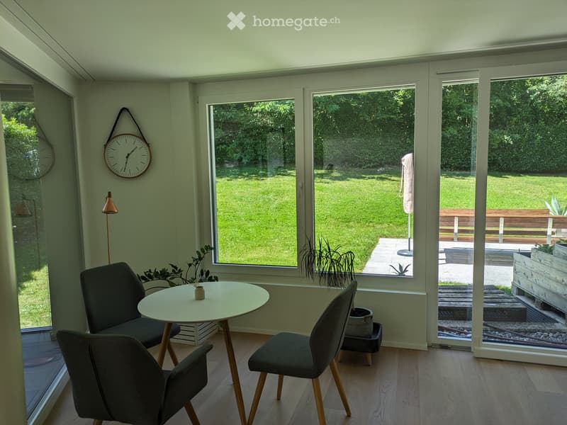 Bright room with large windows overlooking a garden, featuring a round table, chairs, a clock, and a floor lamp.