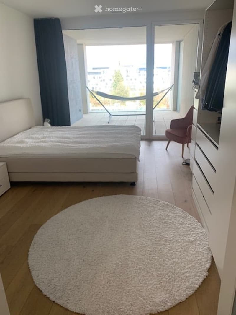 Modern bedroom with a large bed, round shag rug, a chair, and a wardrobe. A hammock is visible on the balcony.