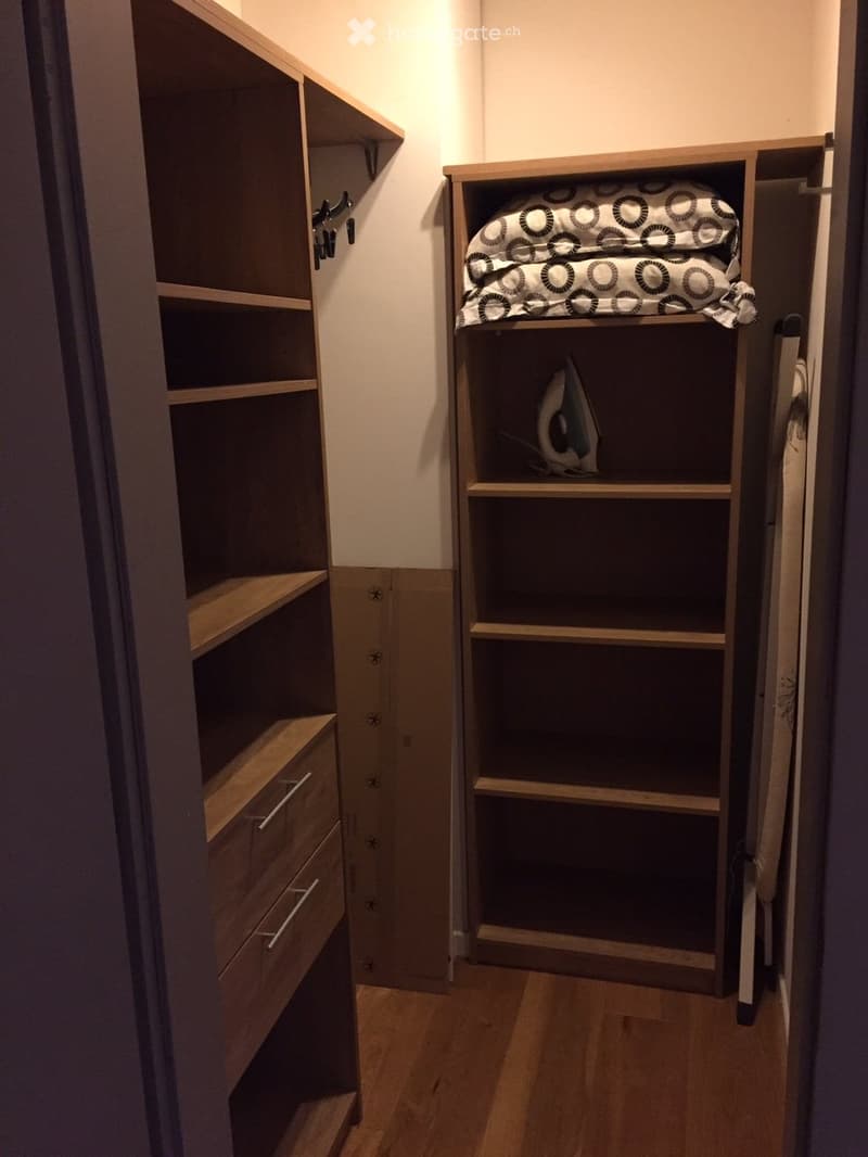 A small, well-organized closet with built-in wooden shelves and drawers, containing an ironing board and iron, and a folded blanket on a shelf.