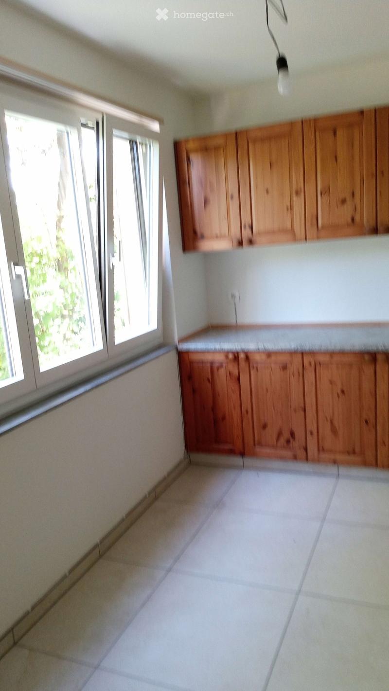 Empty kitchen with wooden cabinets, white tiled floor, and large windows letting in natural light.