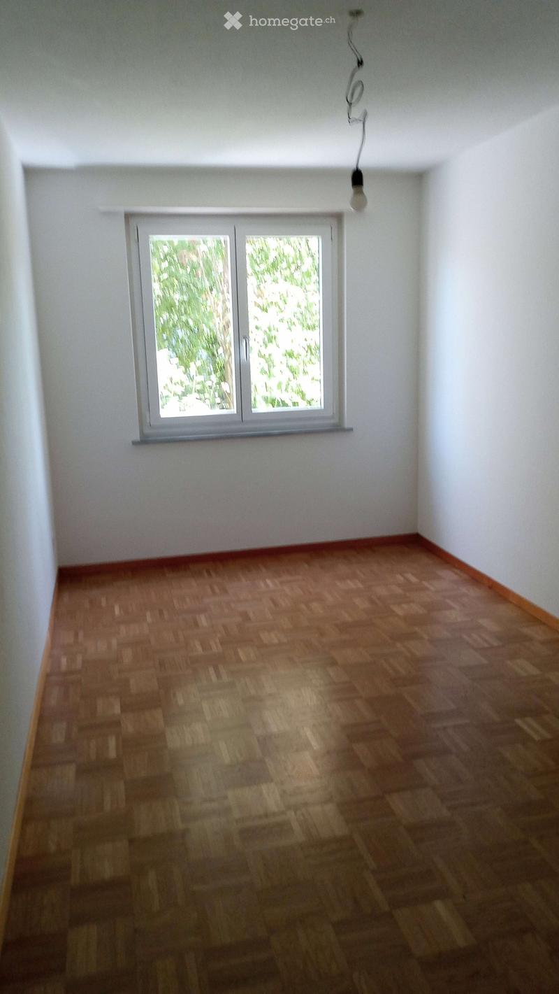 Empty room with wooden flooring and white walls, featuring a large window with a view of greenery outside.