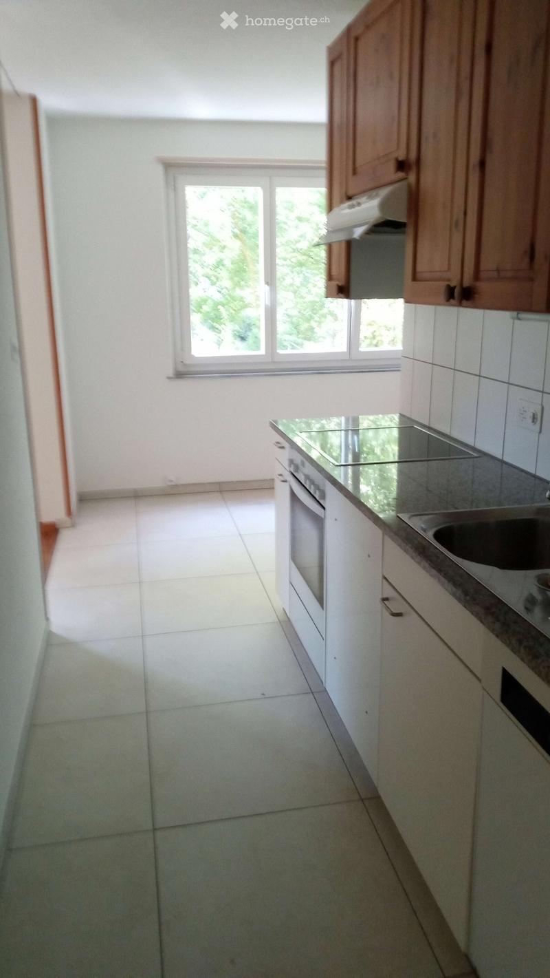 Empty kitchen with white tiled floor, white cabinets, and a window overlooking greenery. No appliances or furniture are present.