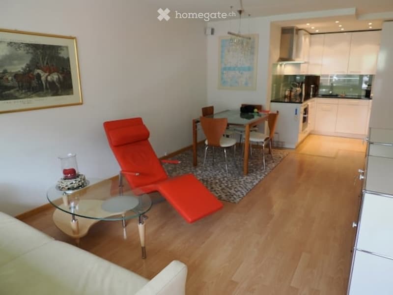 Open-plan living and dining area with a red lounge chair, glass coffee table, sofa, dining table with chairs, and a modern kitchen with white cabinets and stainless steel appliances.