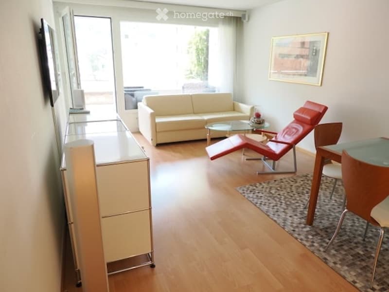 Modern living room with a white sofa, red chair, glass coffee table, and a small dining area with a table and chairs. There is a large window with a view of a balcony.