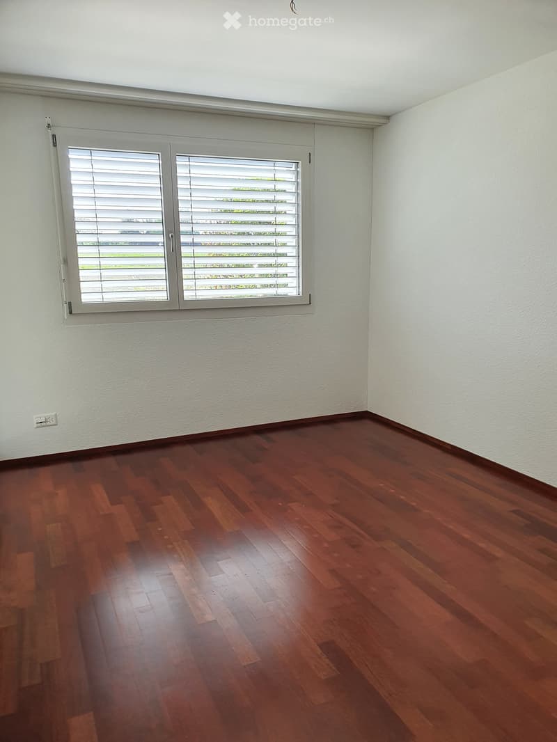 Empty room with wooden flooring and white walls, featuring a large window with blinds.