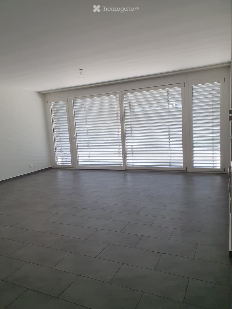 Empty room with grey tiled floor, white walls, large sliding glass doors with blinds, and a single light fixture on the ceiling.