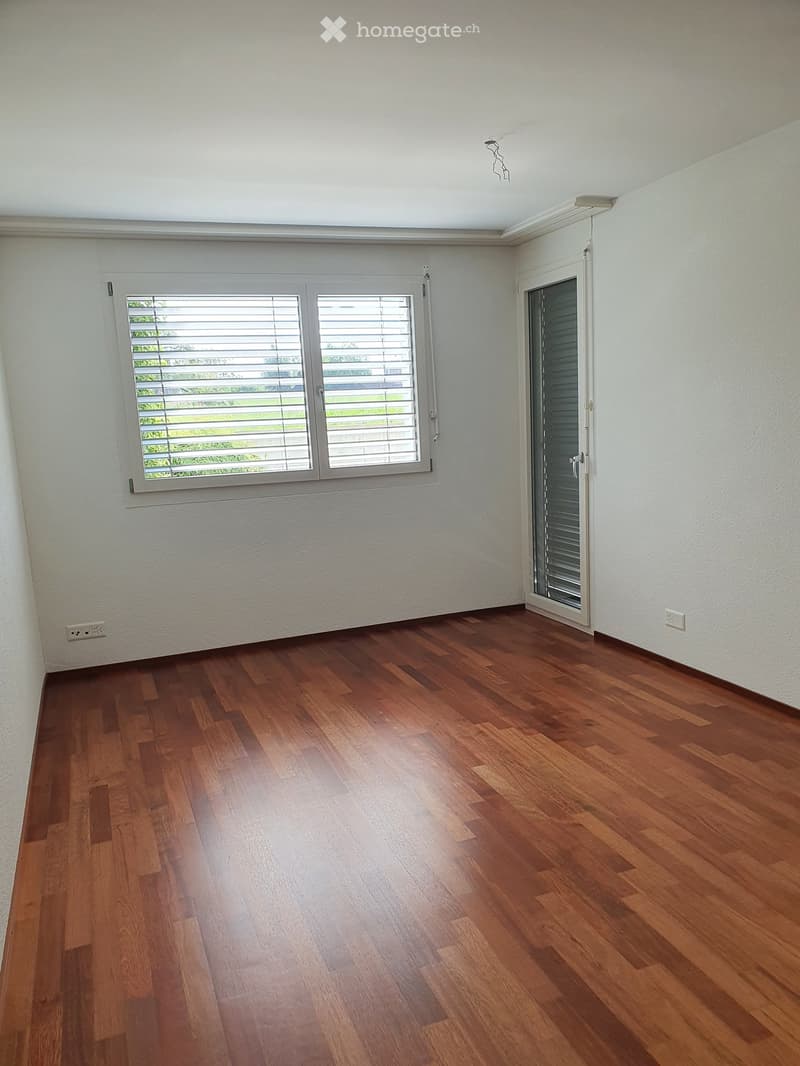 Empty room with wooden flooring, white walls, large window with blinds, and a glass door leading outside.