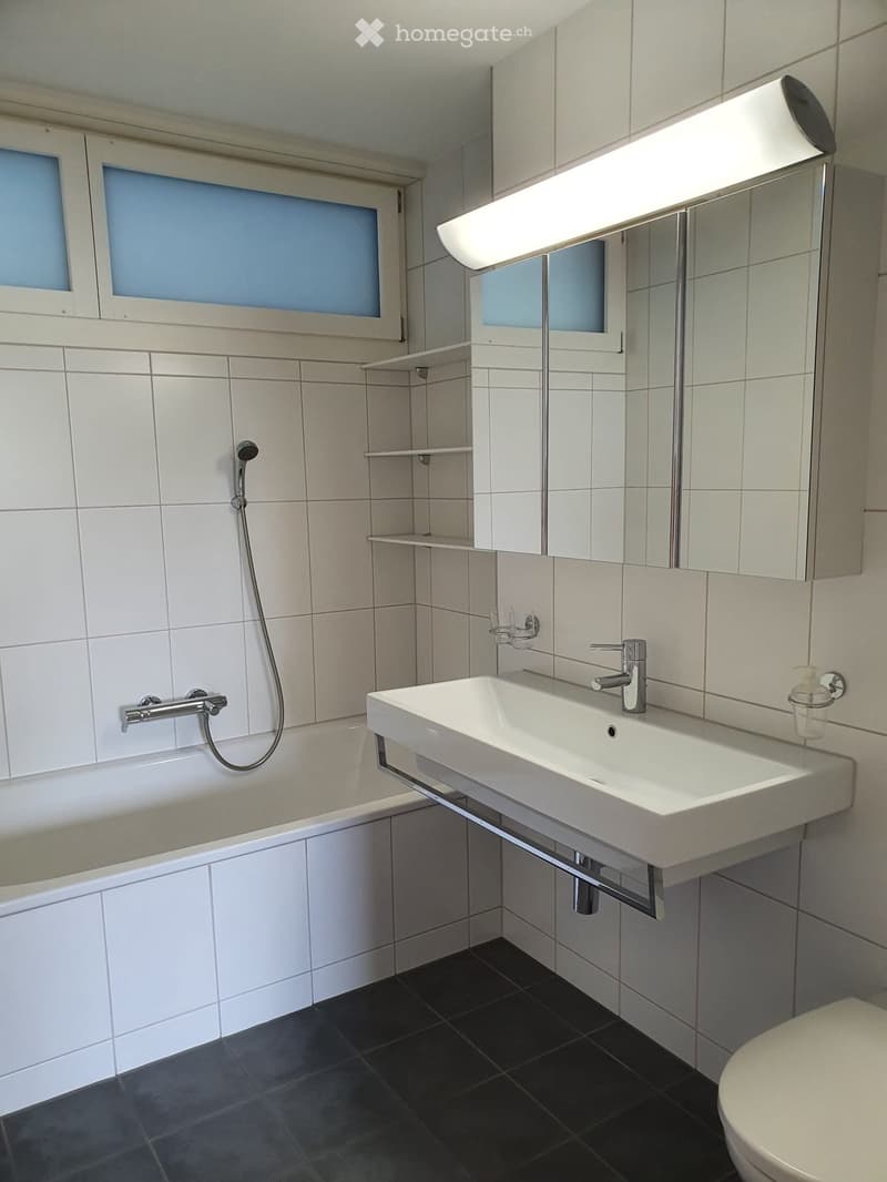 Modern bathroom with white tiles, a bathtub, a sink with a modern faucet, a large mirror with built-in lighting, and a toilet. The walls are tiled in light gray, and the floor is dark gray.