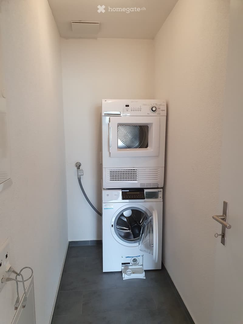 Compact bathroom with a stacked washing machine and dryer unit, white walls, dark tiled floor, and a door visible on the right side.
