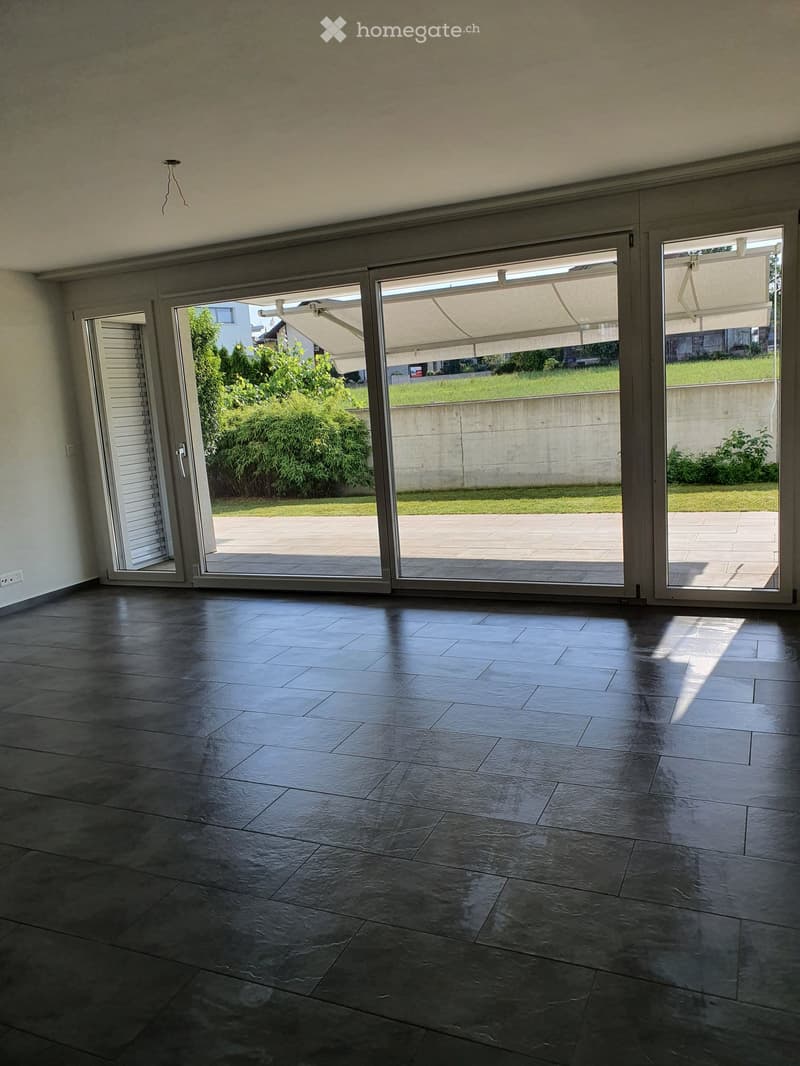 Empty room with dark tiled floor and large sliding glass doors leading to a patio area with a concrete floor and greenery visible outside.