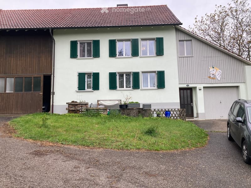 Two-story house with white walls, green shutters, and a red-tiled roof. Features include a wooden garage door, a small garden with a bench, and a driveway with a parked car.